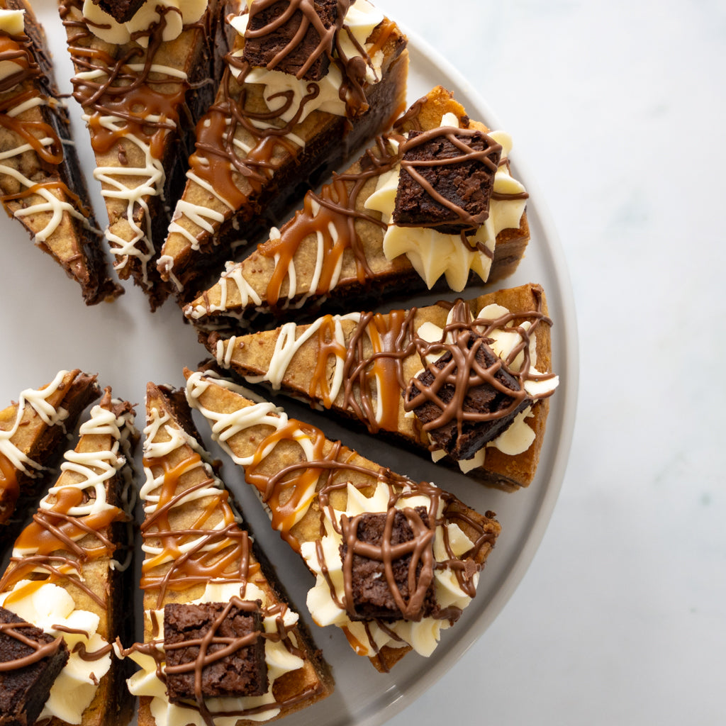 Slices of a layered dessert cookie pie on a white plate, featuring caramel brownie layers, white cream, caramel drizzle, and chocolate brownie squares on a light marble surface.