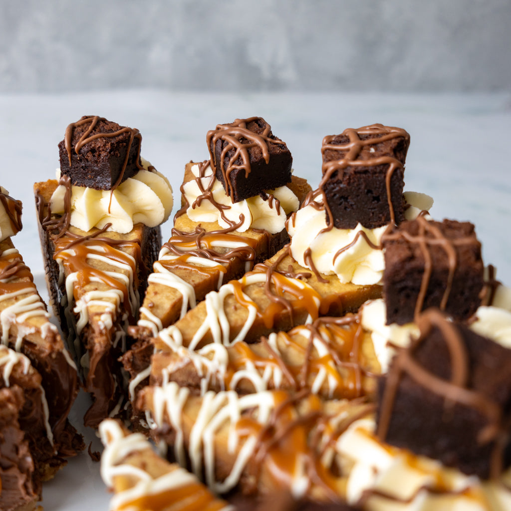 Close-up of several chocolate dessert cookie pie slices topped with whipped cream, banana pieces, drizzles of white and caramel sauce, and a brownie cube on top, arranged on a light surface against a soft grey background.