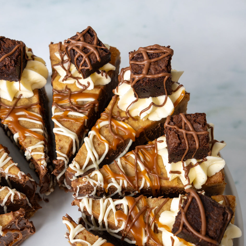 Close-up of several dessert cookie pie slices stacked on a plate, featuring brownie pie with whipped cream, caramel drizzle, and chocolate brownie cubes on top, set against a light marble background.