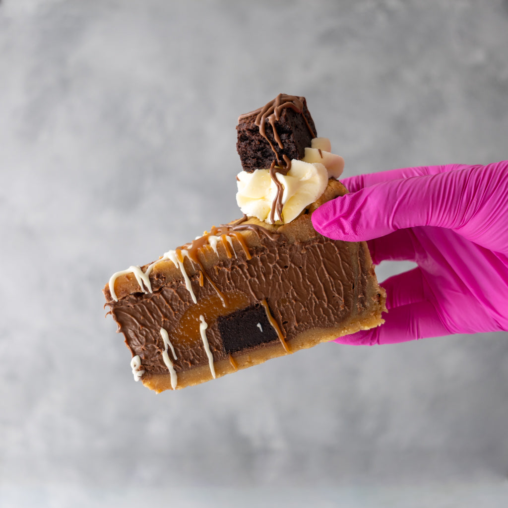 A close-up of a hand wearing a bright pink glove holding a Caramel Brownie Cookie Pie Slice with chocolate frosting, caramel drizzle, whipped cream, and a brownie on top, set against a textured grey background.