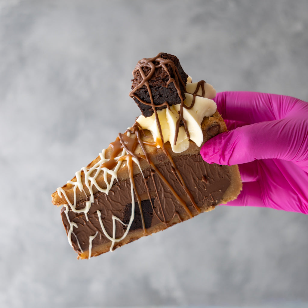 A close-up of a hand wearing a bright pink glove holding a slice of chocolate cookie pie decorated with whipped cream, caramel drizzle, and a brownie on a gray background.