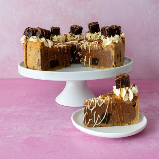 A slice of Full Caramel Brownie Cookie Pie with caramel and whipped cream sits on a small white plate in front of a whole cookie pie on a white pedestal stand, set against a pink background.