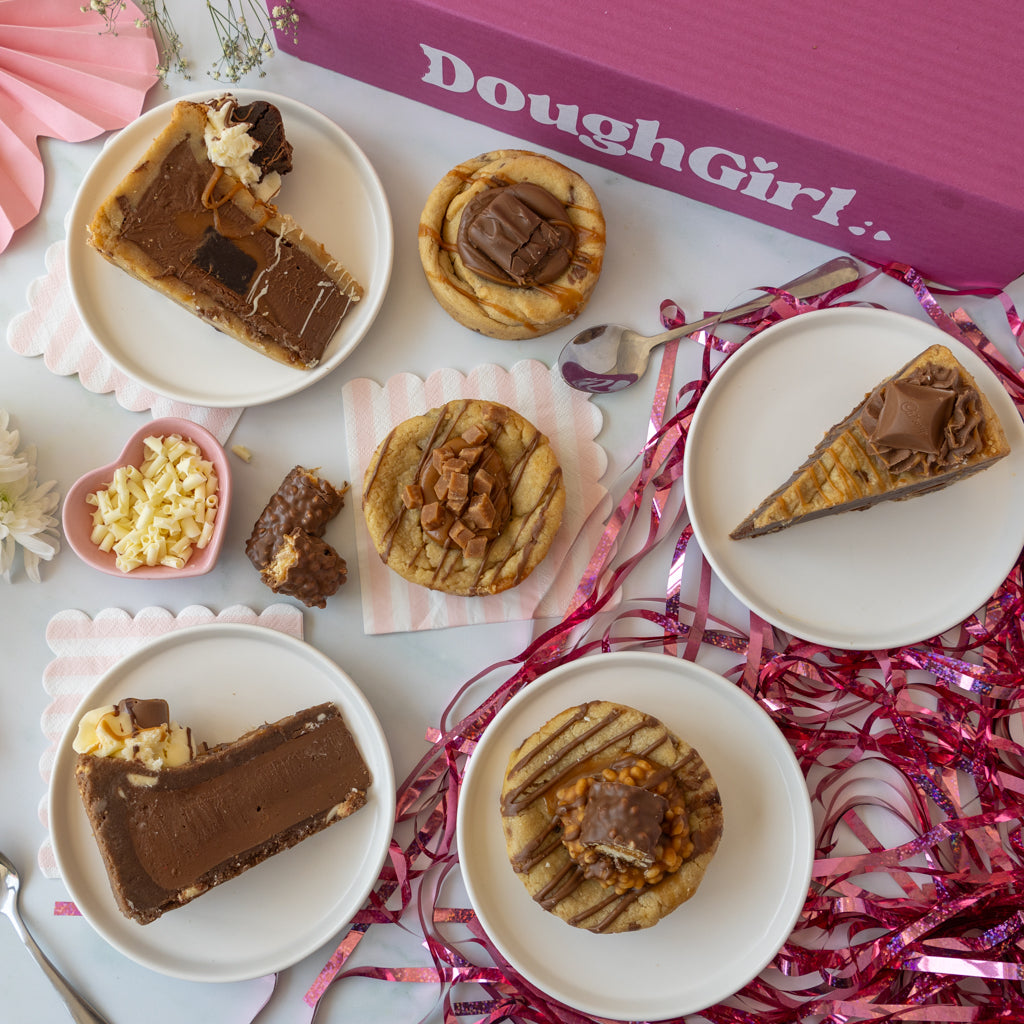 An overhead styled flat-lay of several chocolate-topped baked desserts on white plates, with a pink DoughGirl box in the background and pink ribbon decorations around the scene.