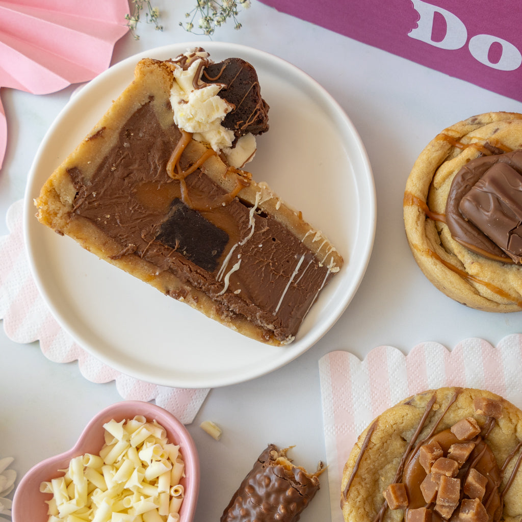 A slice of chocolate-filled pastry on a white plate, surrounded by other desserts including a swirled cookie with chocolate topping, a heart-shaped bowl of shredded white chocolate, and a broken piece of chocolate bar, styled on a light tabletop with pink napkins.