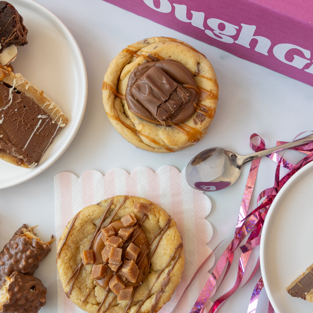 Two chocolate-topped cookies arranged on a white surface with dessert slices, a spoon, pink ribbon, and part of a pink branded box visible in the background.