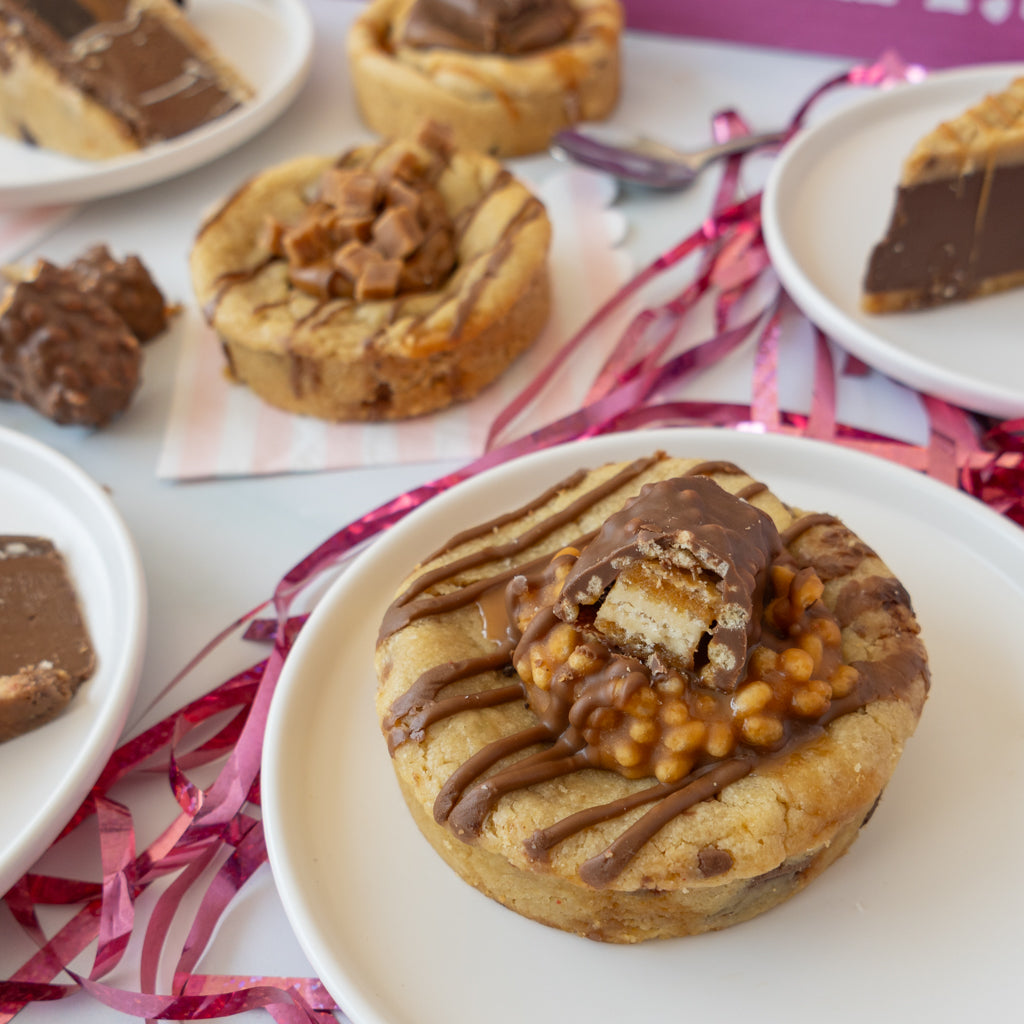 A close-up of a chocolate-topped cookie on a white plate, with other similar desserts and pink ribbon scattered around on a white surface.