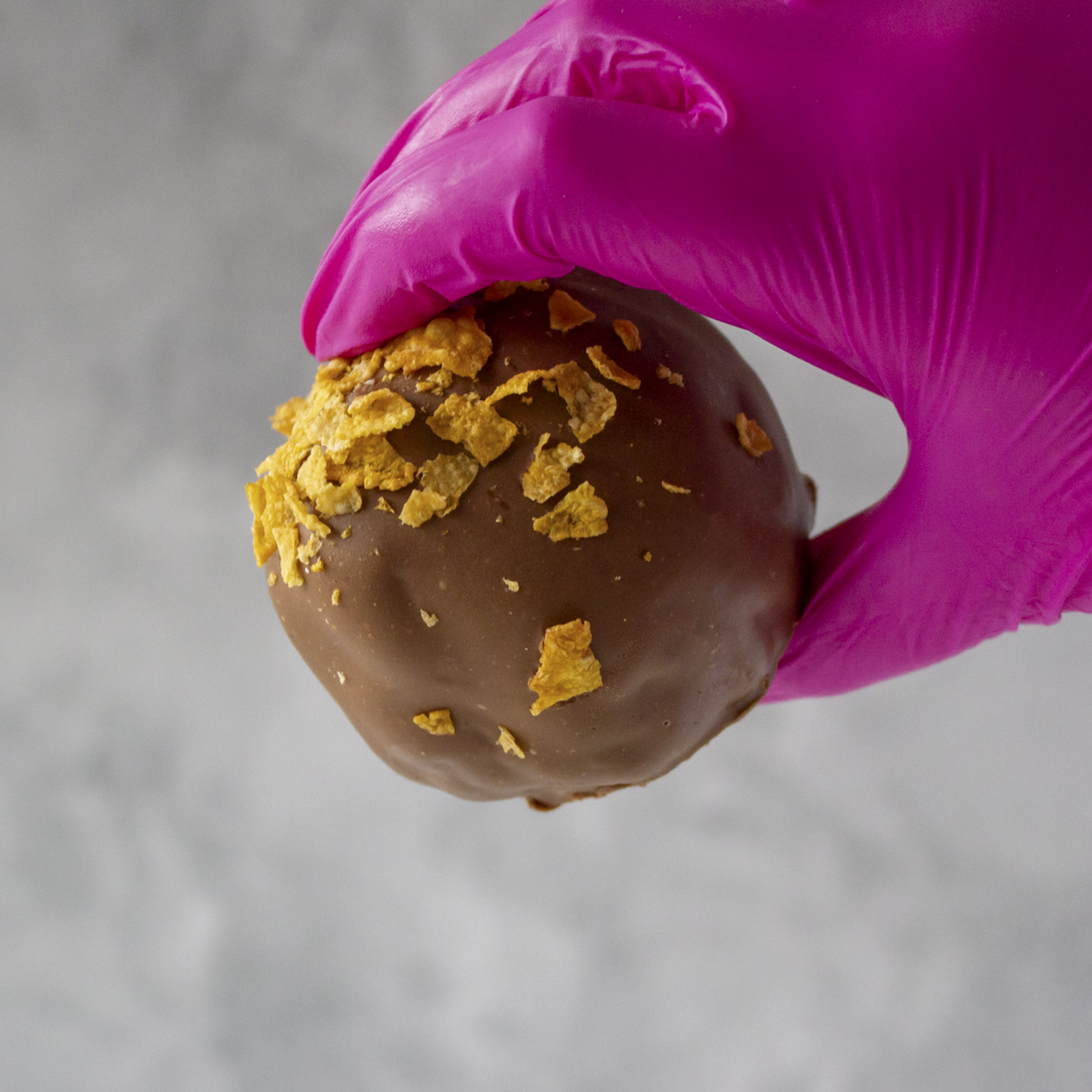 Caramel Cornflake Cracker Choccy Boom Bites with cornflake pieces held by a hand in a pink glove against a gray background