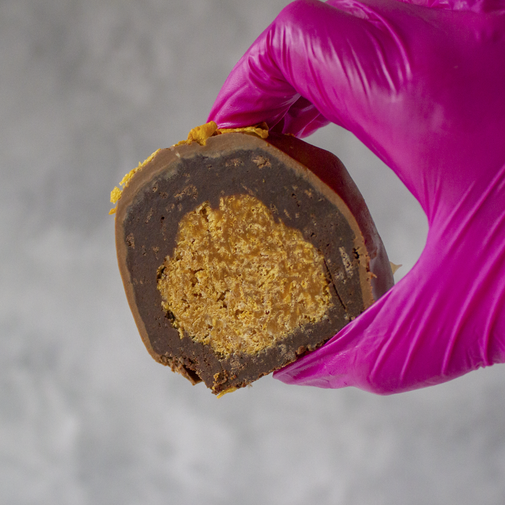 Caramel Cornflake Cracker Choccy Boom Bites by DoughGirl UK with a golden caramel cornflake centre held by a hand in a purple glove against a gray background