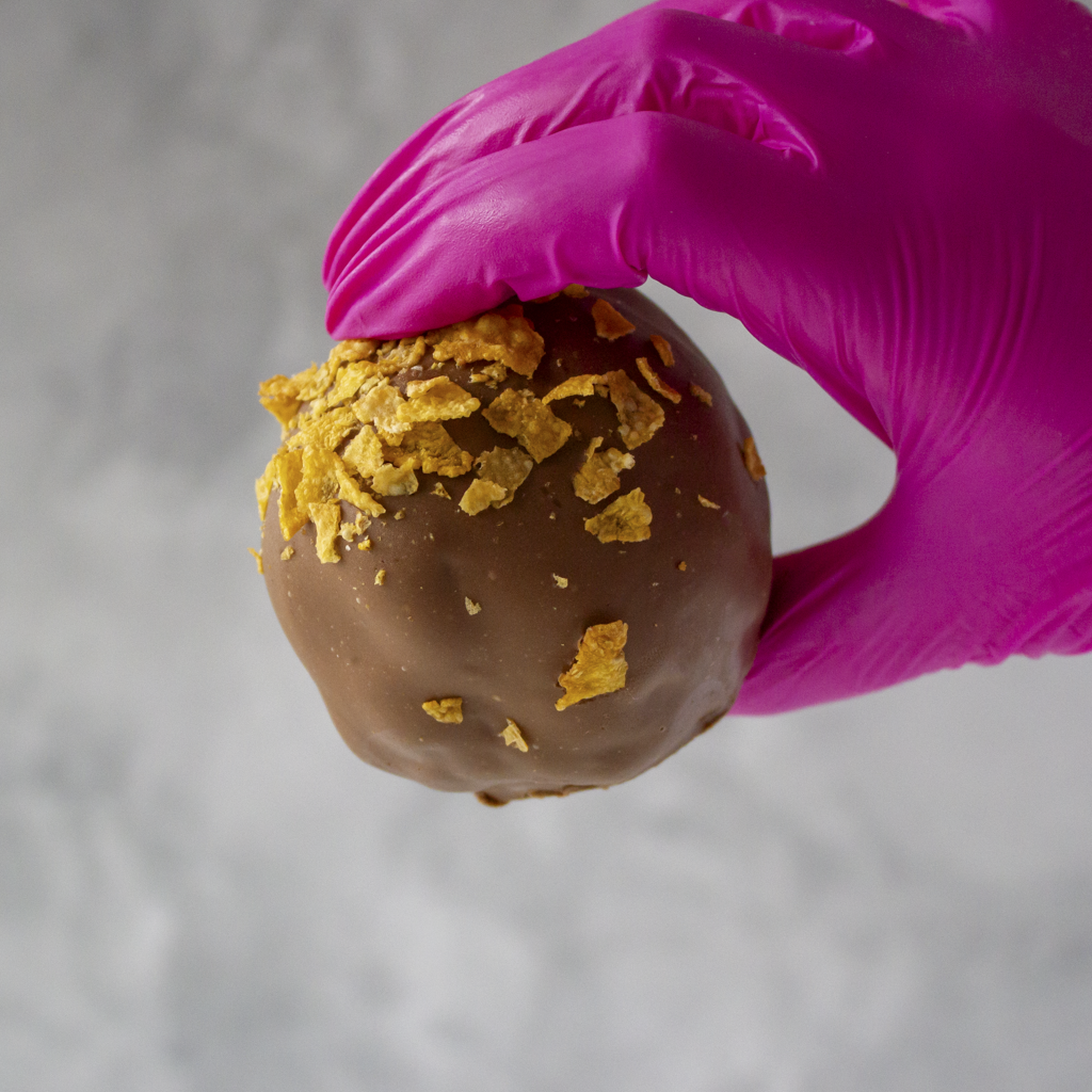 Chocolate-covered treat with cornflake pieces held by a hand in a pink glove on a gray background