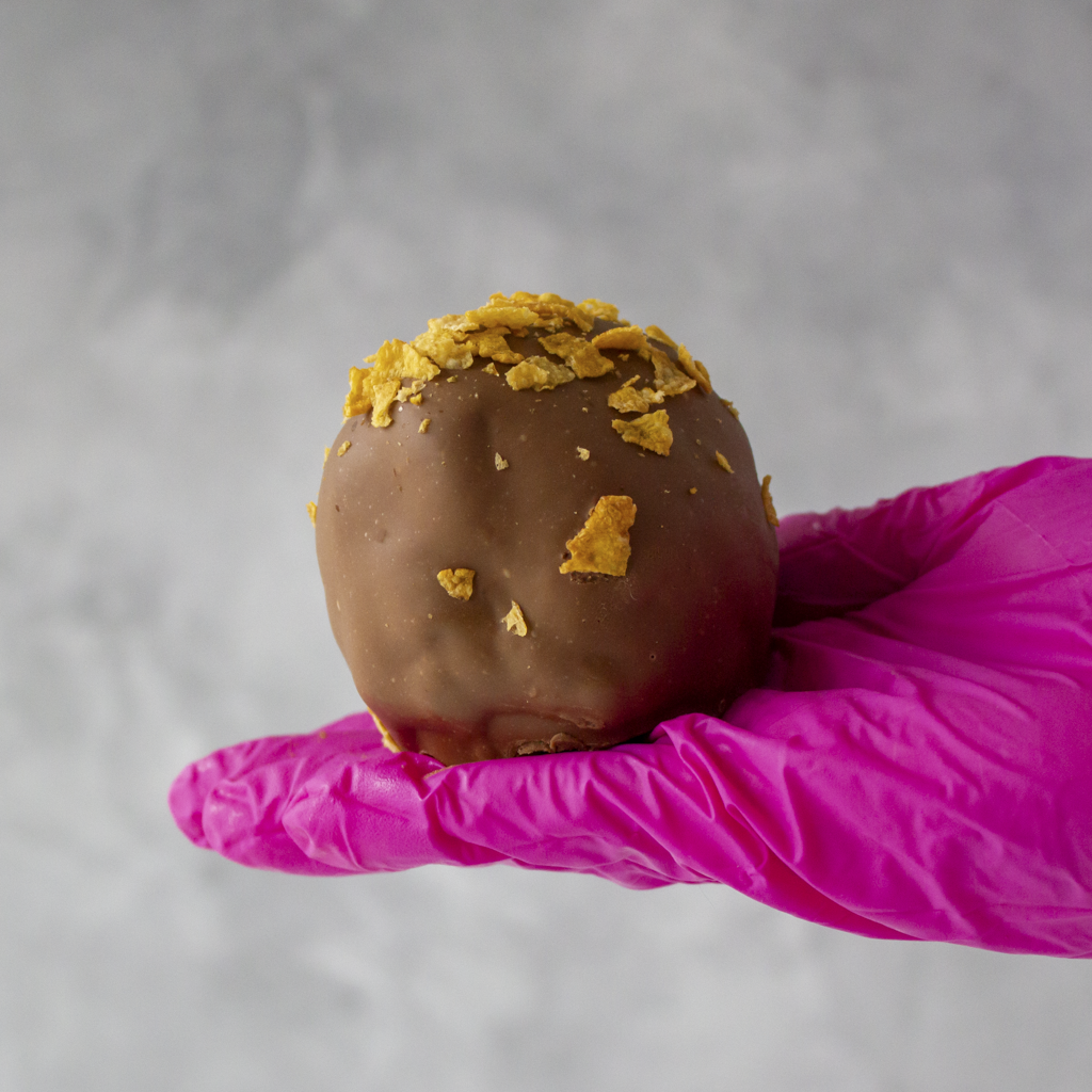 Caramel Cornflake Cracker with cereal pieces held by a hand wearing a pink glove on a gray background