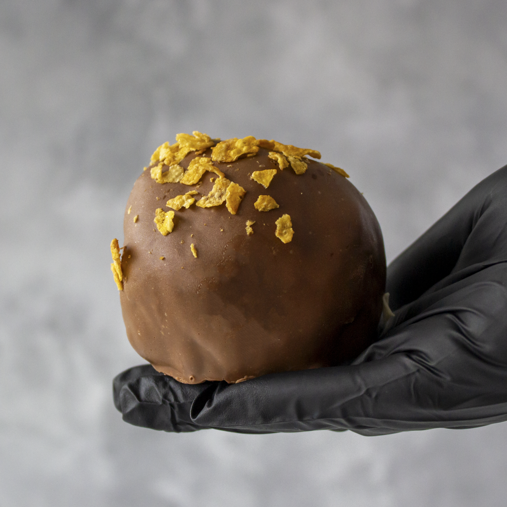 Caramel Cornflake Cracker Choccy Boom Bites with cornflake held by a black gloved hand against a gray background