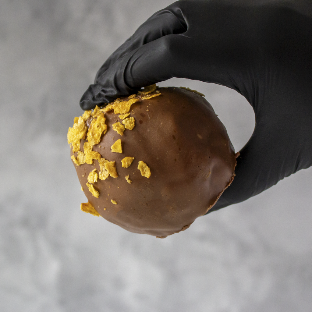 Chocolate-covered dessert with cornflakes held by a gloved hand against a neutral background