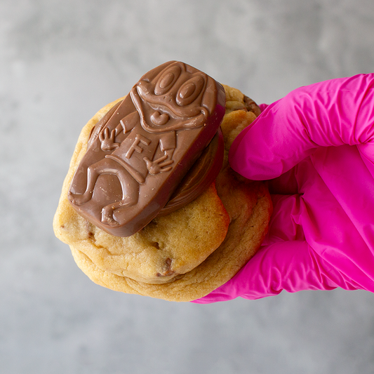 Hand wearing a pink glove holding a Caramel Freddo Deluxe Cookie with a chocolate bar on a gray background