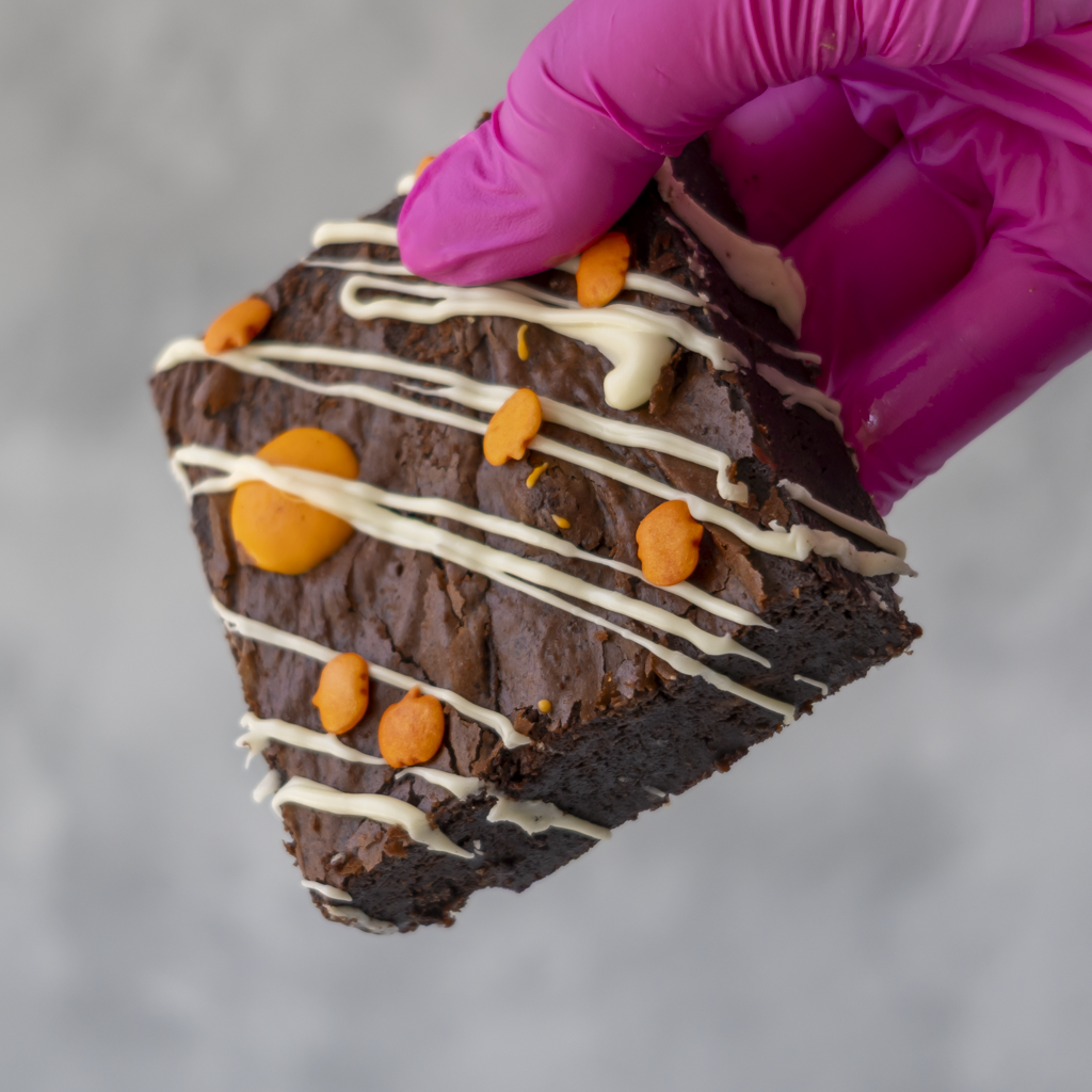 A close-up of a hand wearing a bright pink glove holding a triangular slice of Spellbound Chocolate & Orange Brownie decorated with white chocolate drizzle and orange cookie pieces, against a soft gray background.
