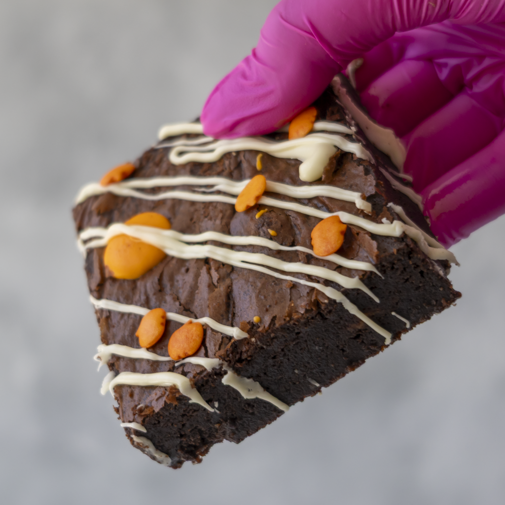 A close-up of a hand wearing a bright pink glove holding a triangular slice of chocolate brownie decorated with white drizzle and orange chocolate pieces, against a soft gray background.