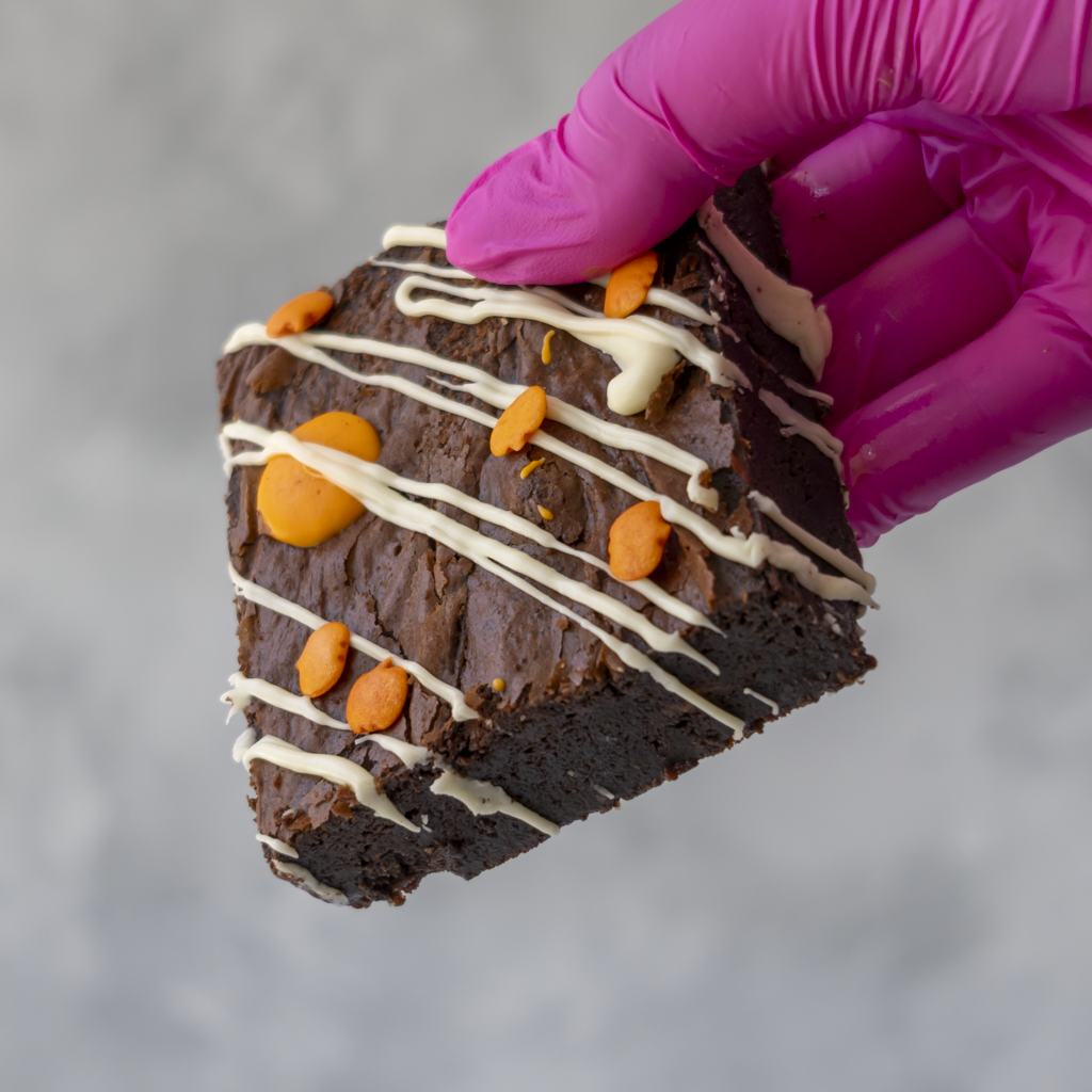A close-up of a hand wearing a bright pink glove holding a triangular slice of chocolate brownie topped with white drizzle, and orange chocolate crumbs, against a soft gray background.
