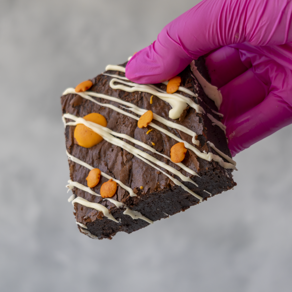 A hand wearing a bright pink glove holds a square brownie slab topped with white chocolate drizzle, orange chocolate pieces, against a soft grey background.