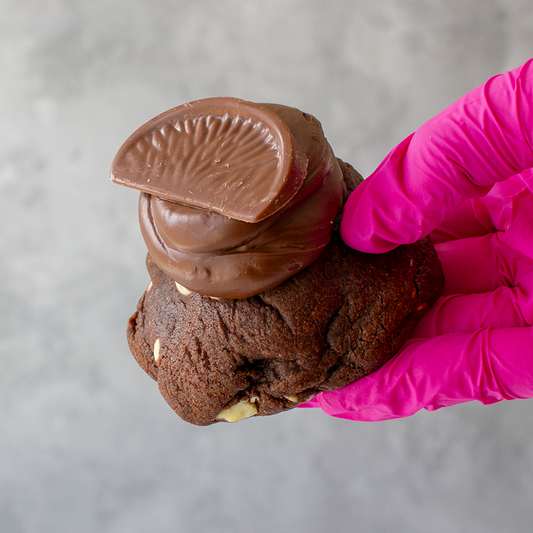Chocolate Orange Deluxe Cookie with a stack of chocolate coins held by a hand wearing a pink glove on a gray background