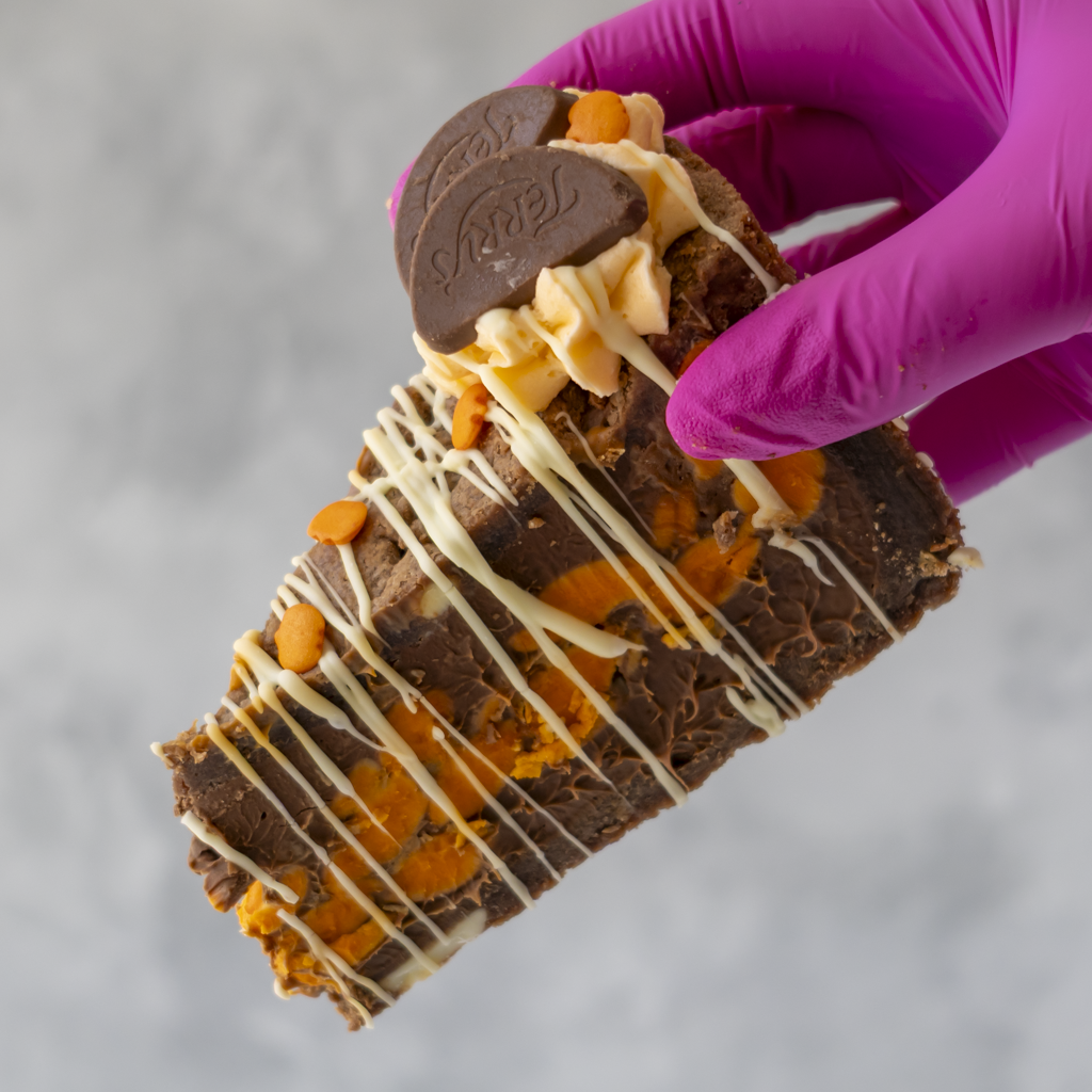 A hand wearing a bright purple glove holds a thick slice of Chocolate & Orange Fright Cookie Pie, white chocolate drizzle, and cookie crumbs against a light grey background.