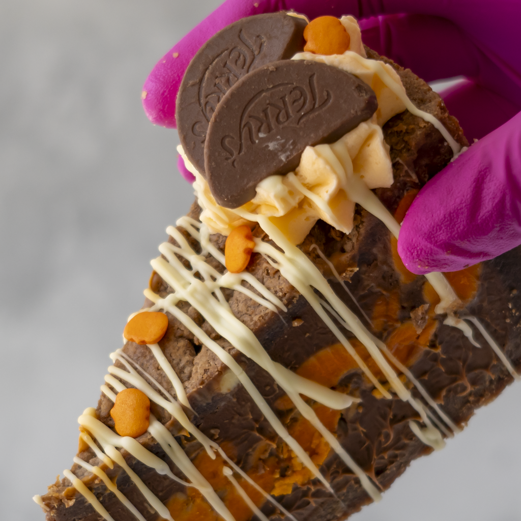 A close-up of a hand wearing a bright pink glove holding a decorated chocolate slice or pastry. The dessert has dark chocolate layers, white chocolate drizzle, orange chocolate filling, and two dark chocolate Oreo-style pieces on top, against a soft light grey background.