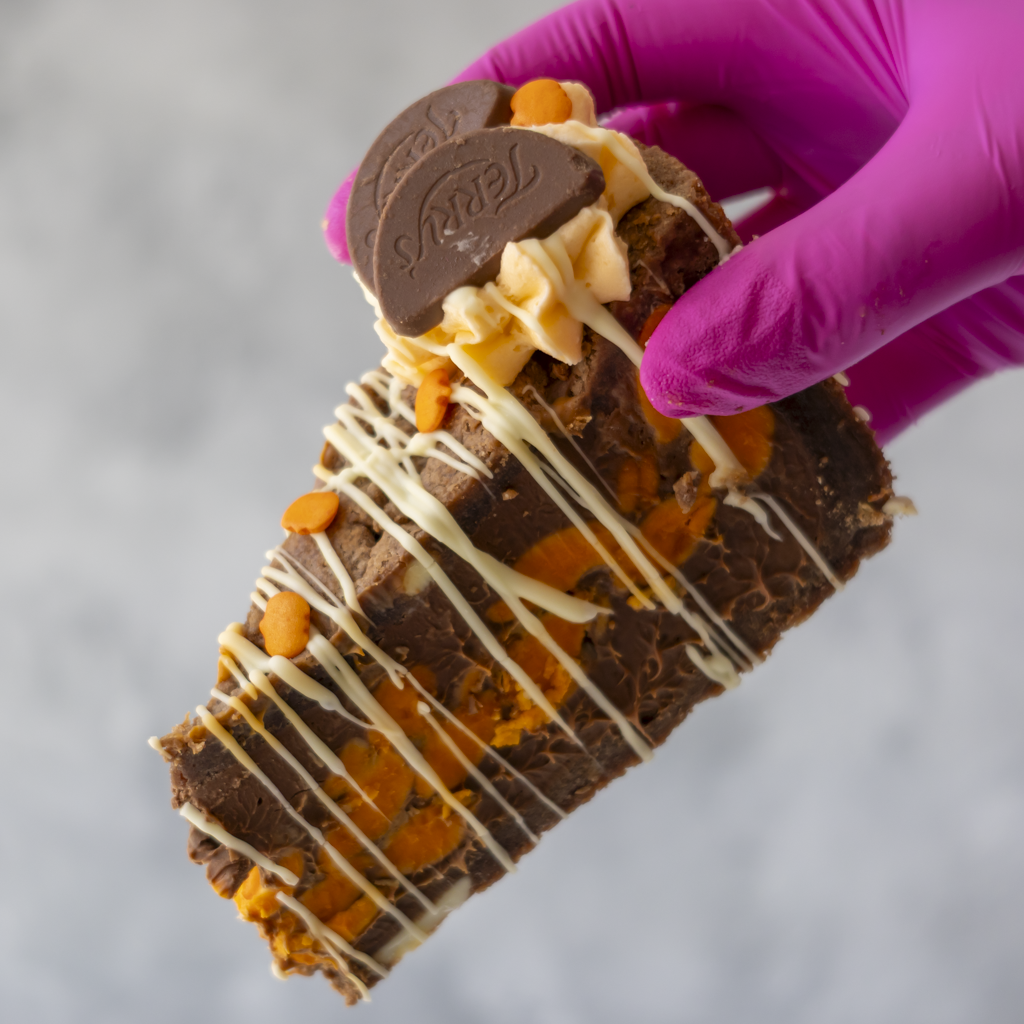 A close-up of a hand wearing a bright pink glove holding a chocolate-covered cookie pie slice with white chocolate drizzle, caramel-like orange chunks, and a dark chocolate coin on top, against a soft light grey background.