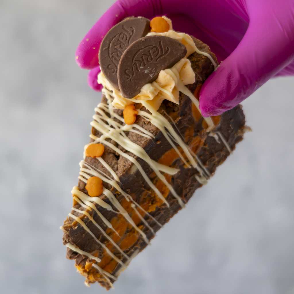 A hand wearing a bright pink glove holds a triangular slice of chocolate-covered cookie pie topped with white chocolate drizzle, cream, and two dark chocolate Terry pieces, against a soft grey background.