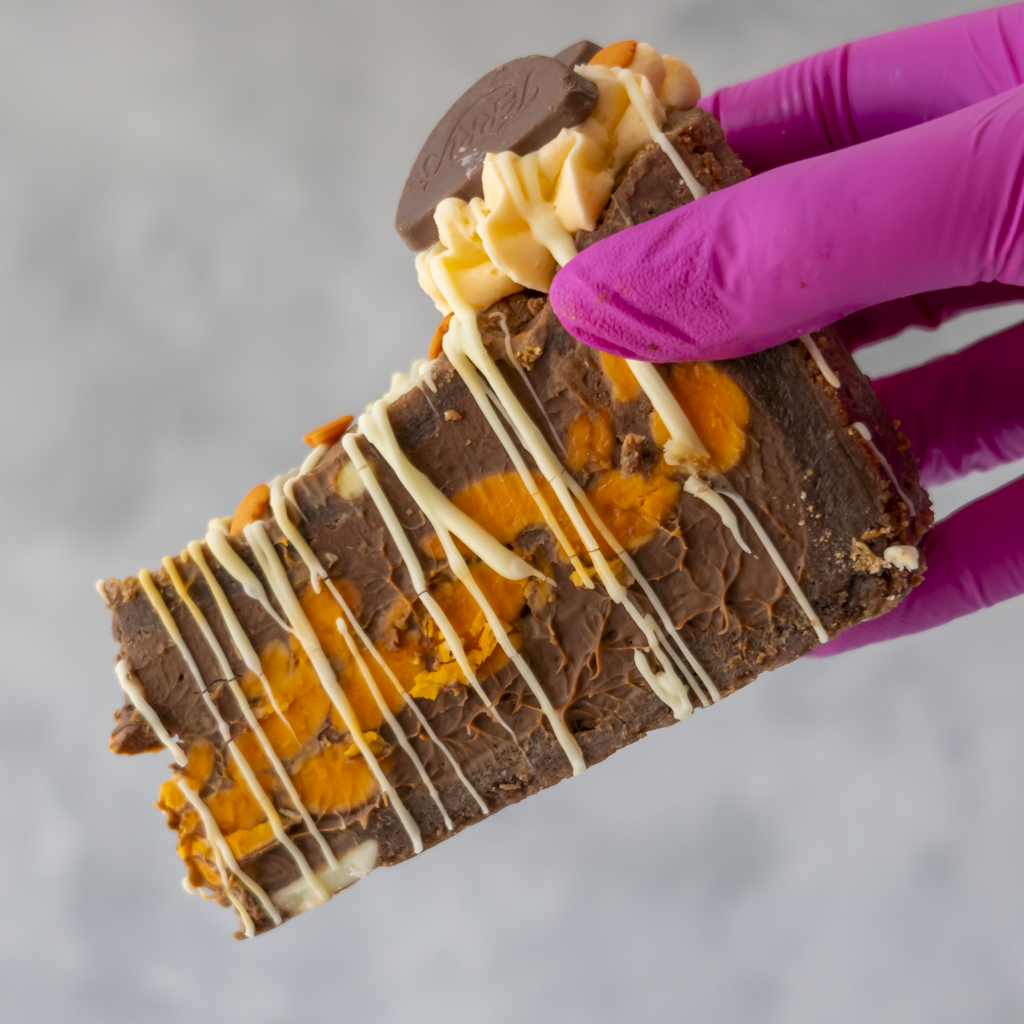 A hand wearing a bright magenta glove holds a thick slice of chocolate bark decorated with drizzles of white chocolate, orange buttercream, and orange pieces, against a soft gray background.