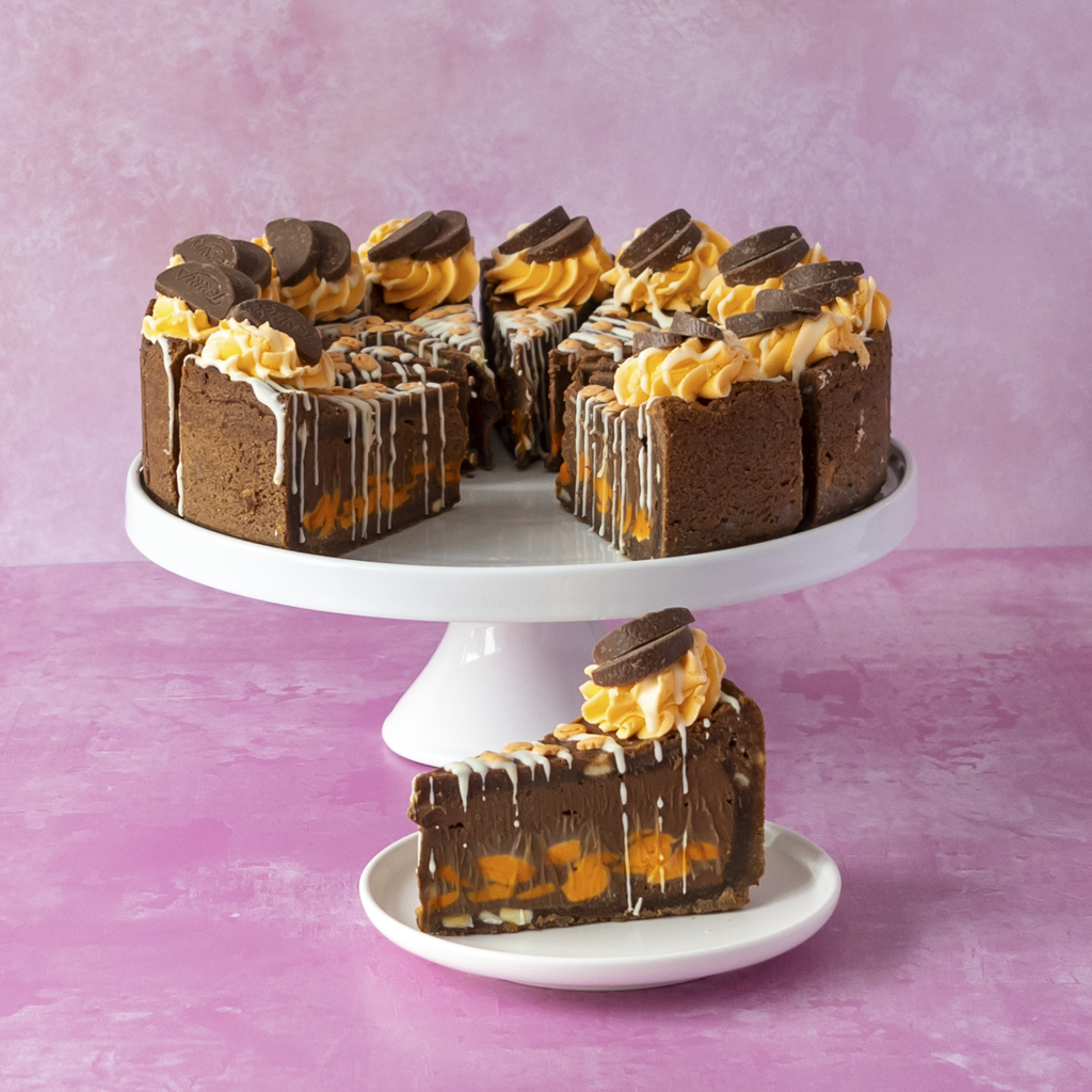 Chocolate & Orange Fright Cookie Pie on a white cake stand with a single slice on a small plate in front, set against a pink background. The cake has orange chocolate filling and is topped with chocolate cookie pieces and white drizzle.