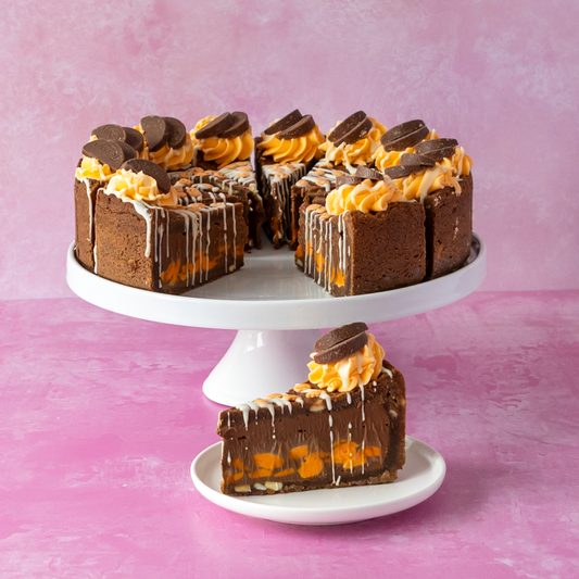 Chocolate & Orange Fright Cookie Pie on a white cake stand with a single slice on a small plate in front, set against a pink background. The cake has orange chocolate filling and is topped with chocolate cookie pieces and white drizzle.