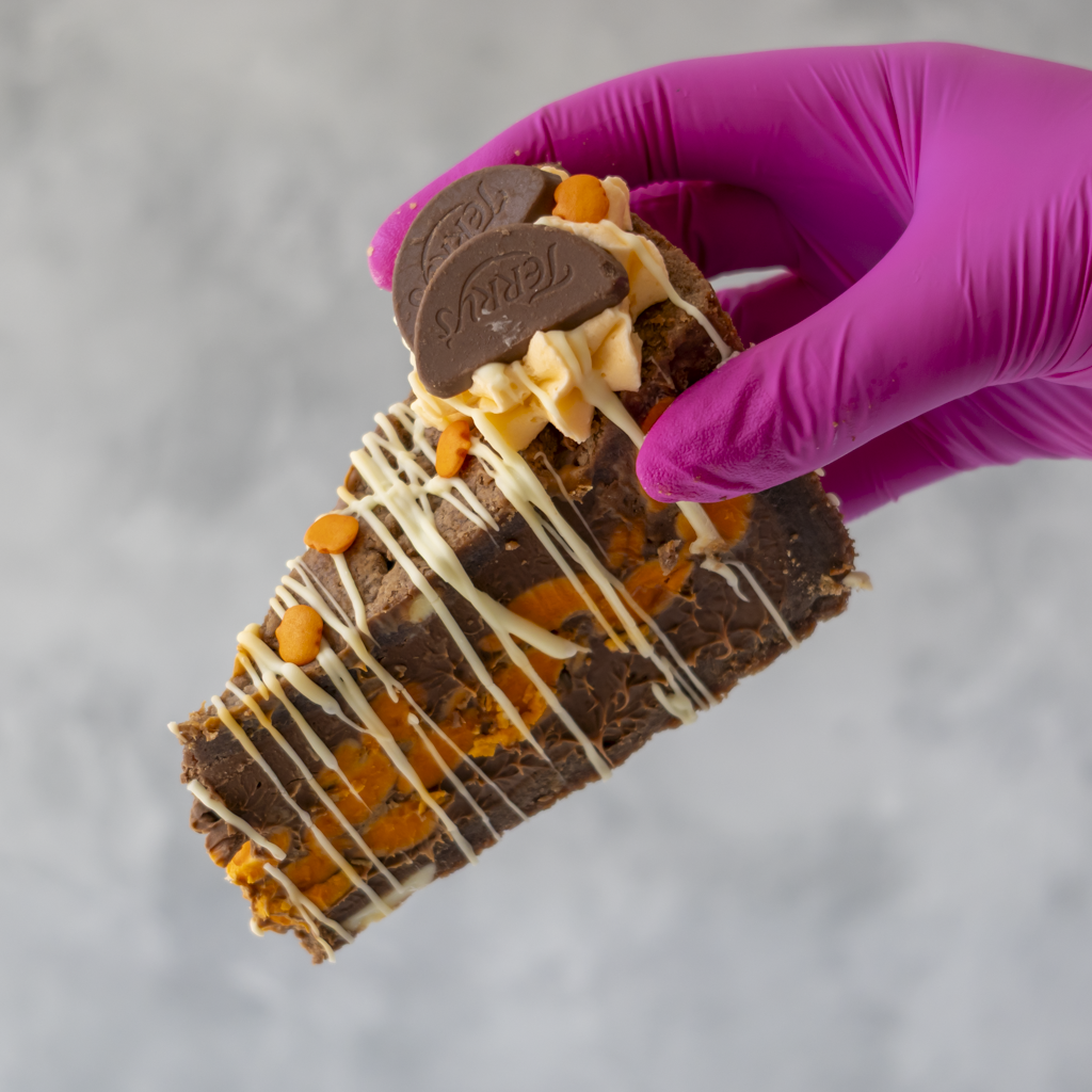 A hand wearing a bright magenta glove holds a triangular slice of Chocolate & Orange Fright Cookie Pie decorated with white drizzle, orange chocolate pieces, and two Terry chocolate discs, against a soft grey background.