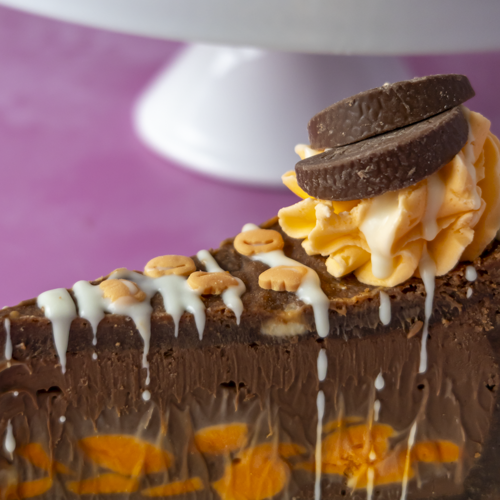 A close-up of a slice of chocolate pie with orange cream swirls, white chocolate drizzle, orange chocolate, and two orange chocolate pieces on top, set against a pink background.