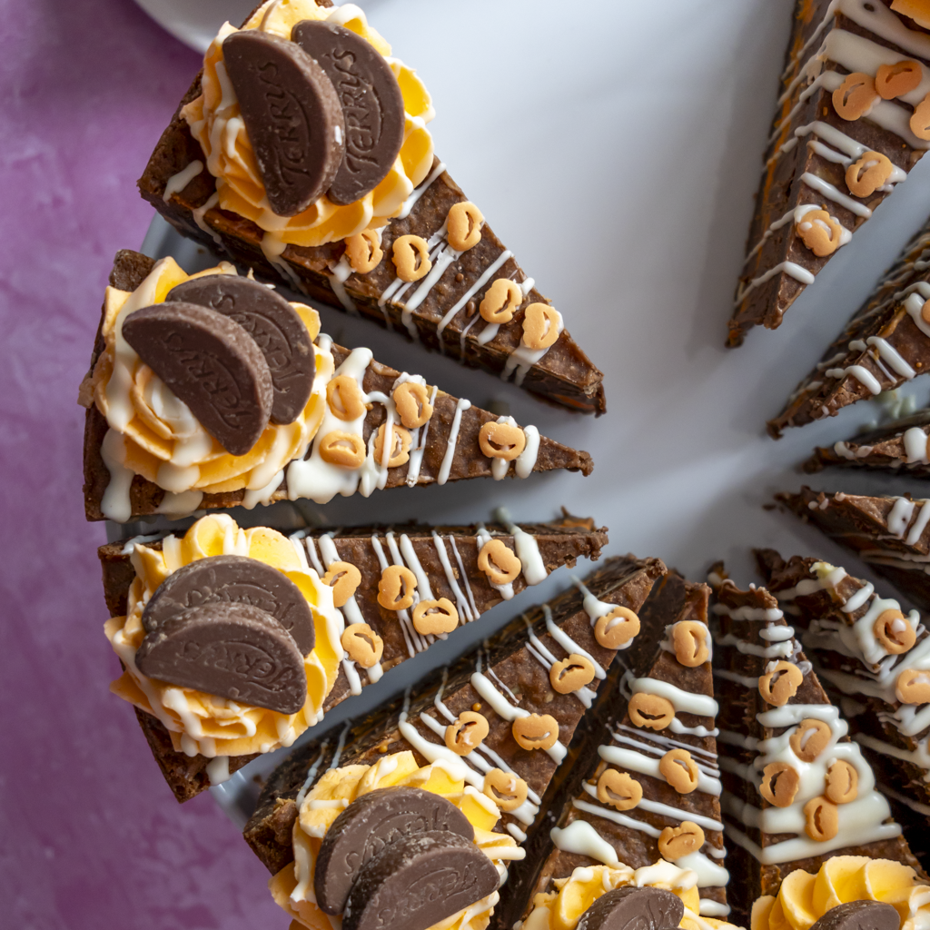 Slices of a chocolate cake decorated with orange cream, white drizzle, sprinkles, and orange chocolate pieces, arranged on a white plate with a purple surface visible at the side.