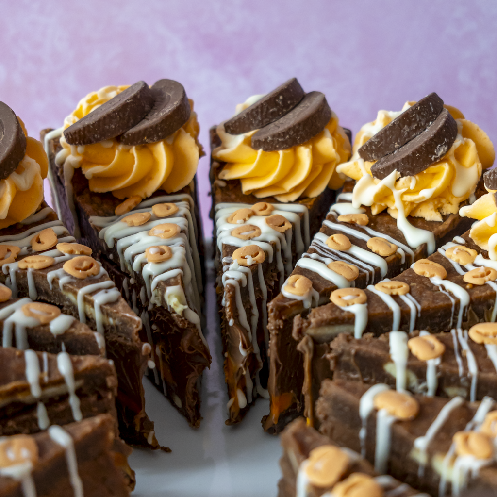 Close-up of several chocolate dessert slices arranged on a plate, decorated with orange cream, white chocolate drizzle, sprinkles, and chocolate slices, against a soft pink background.
