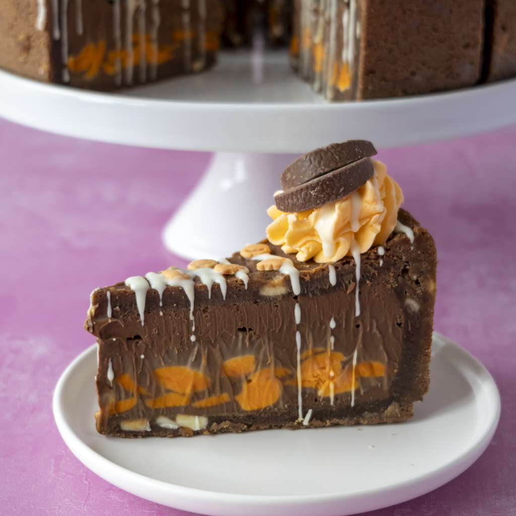 A slice of chocolate pie with orange fruit pieces, white drizzle, and a cookie topping, served on a small white plate with a cake stand in the background.
