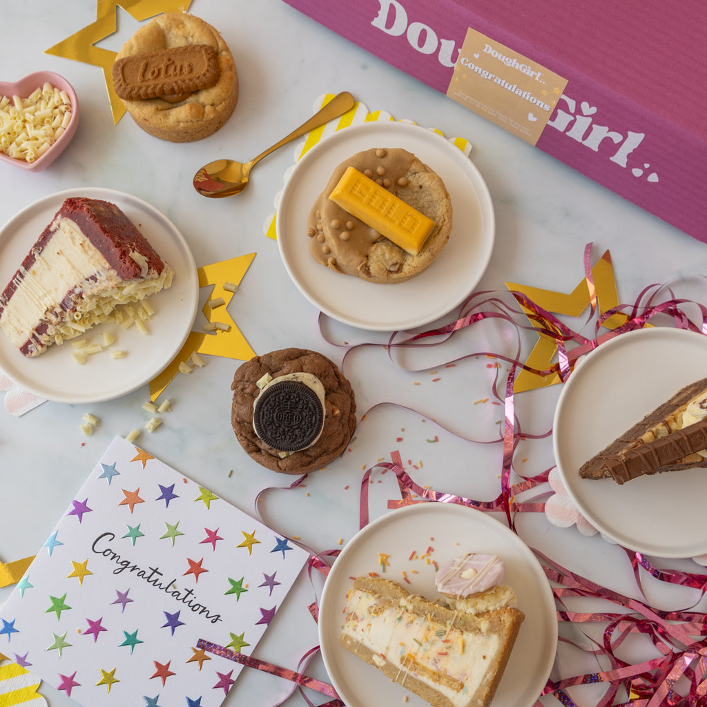 A styled flat-lay of birthday treats and a pink 'DoughGirl' gift box on a white surface, surrounded by plates of cookie pie slices, cookies with chocolate and caramel toppings, a small bowl of shredded cheese, a gold spoon, star-shaped decorations, and a colorful congratulations card.