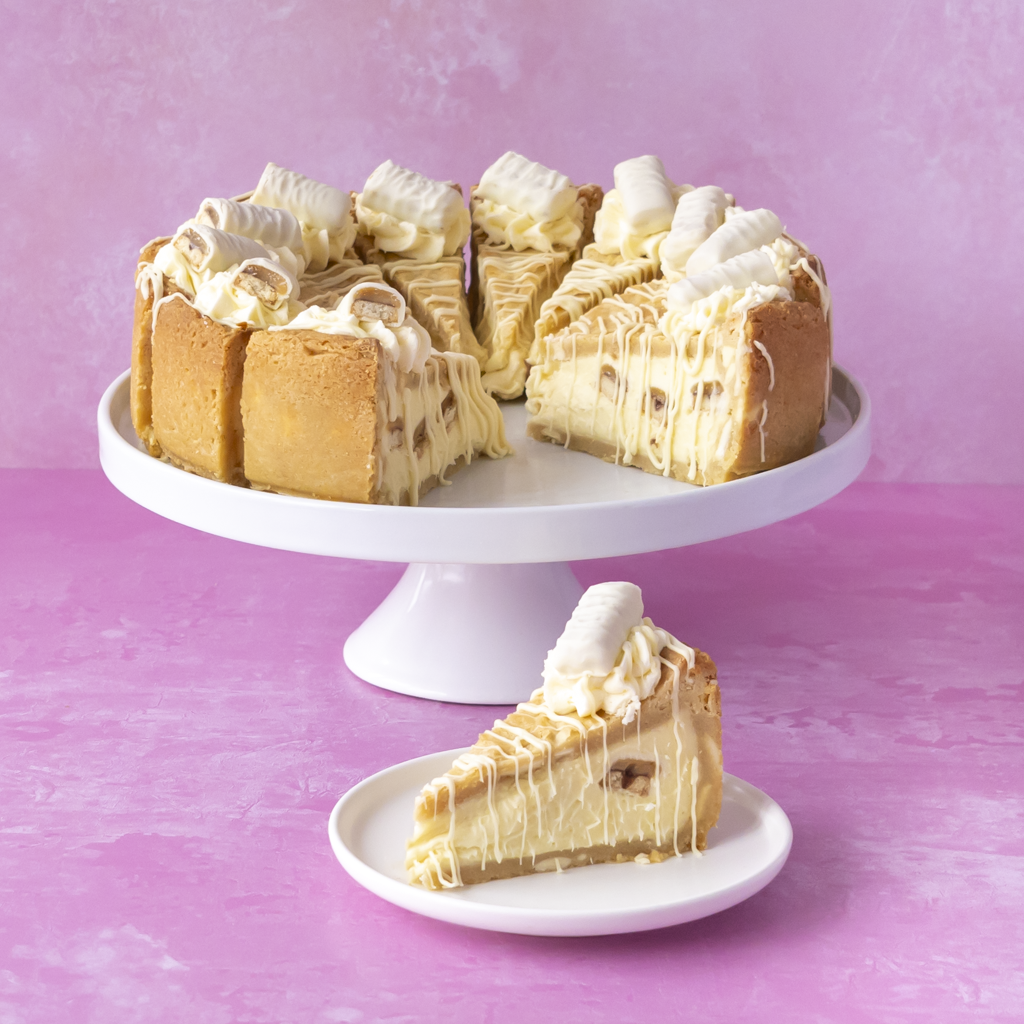 A Cookie Pie with White Twix with whipped cream and white drizzle sits on a white cake stand, with one slice placed on a small matching plate in front, against a pink studio background.