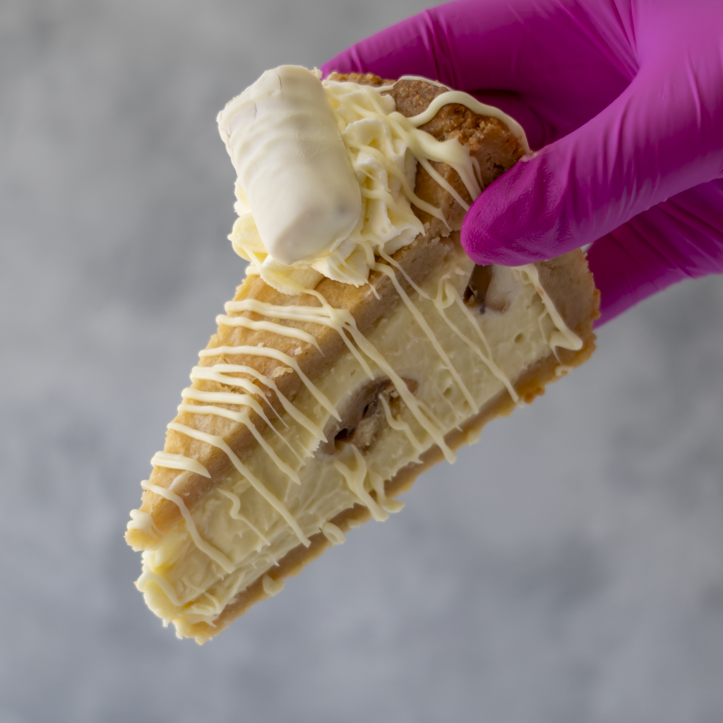 A close-up of a hand wearing a bright pink glove holding a slice of Cookie Pie with White Twix with white chocolate drizzle against a soft gray background.