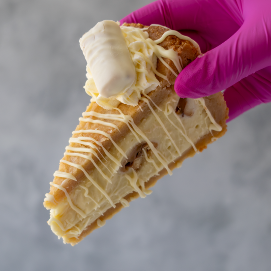 A close-up of a hand wearing a bright pink glove holding a slice of Cookie Pie with White Twix with white chocolate drizzle against a soft gray background.