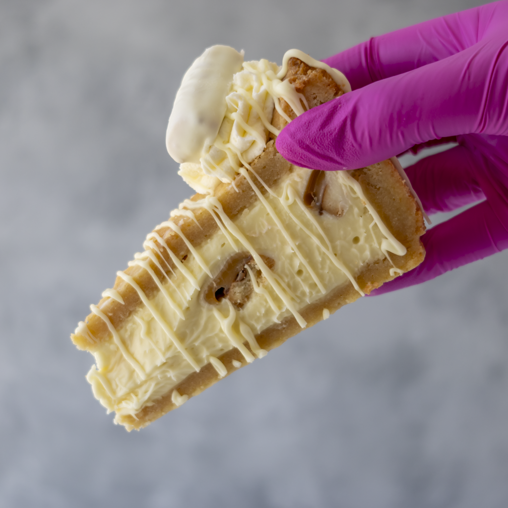 A close-up of a hand wearing a bright magenta glove holding a slice of Cookie Pie with White Twix with white chocolate drizzle and a dollop of white cream, against a soft gray background.