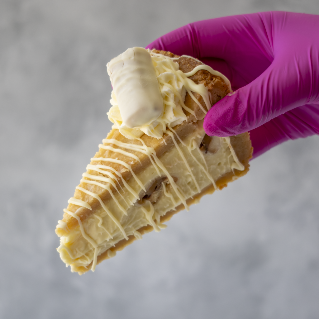 A hand wearing a bright magenta glove holds a Cookie Pie Slice with White Twix with white chocolate drizzle against a soft gray background.