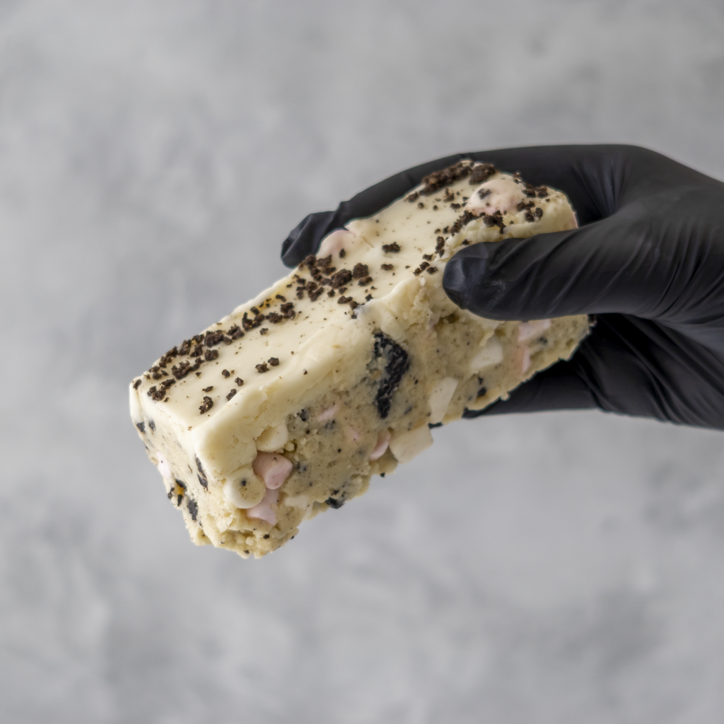 Hand holding a block of Cookies and Cream Rocky Mountain Road cake with chocolate chips and other ingredients against a gray background