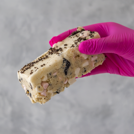 Hand holding a Cookies and Cream Rocky Mountain Road dough with chocolate chips and pink sprinkles against a gray background.