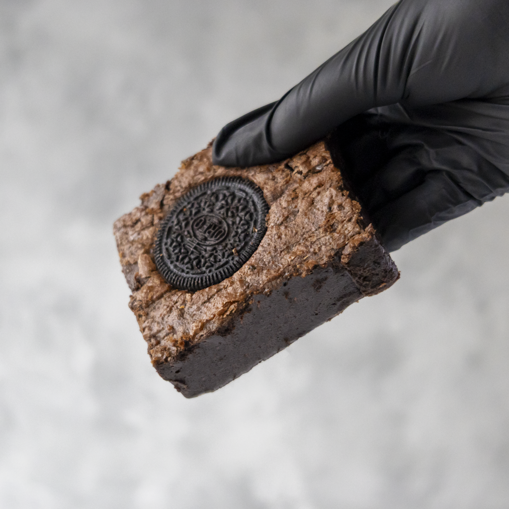 Cookies 'n Cream Brownie Slab packed full of crushed Oreo cookies, held by a gloved hand against a neutral background