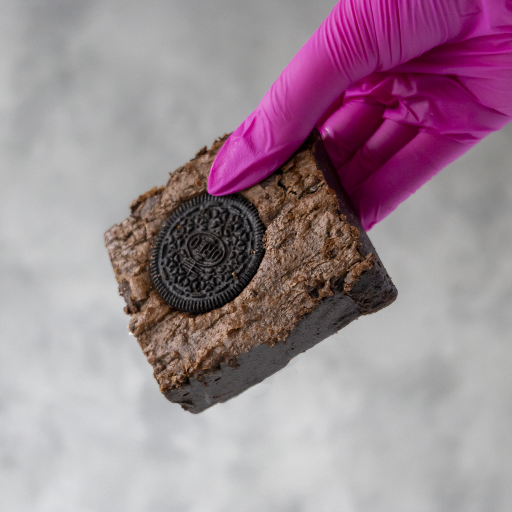 Cookies 'n Cream Brownie Slab with chocolate brownie base and an Oreo cookie piece held by a hand in a pink glove on a neutral background