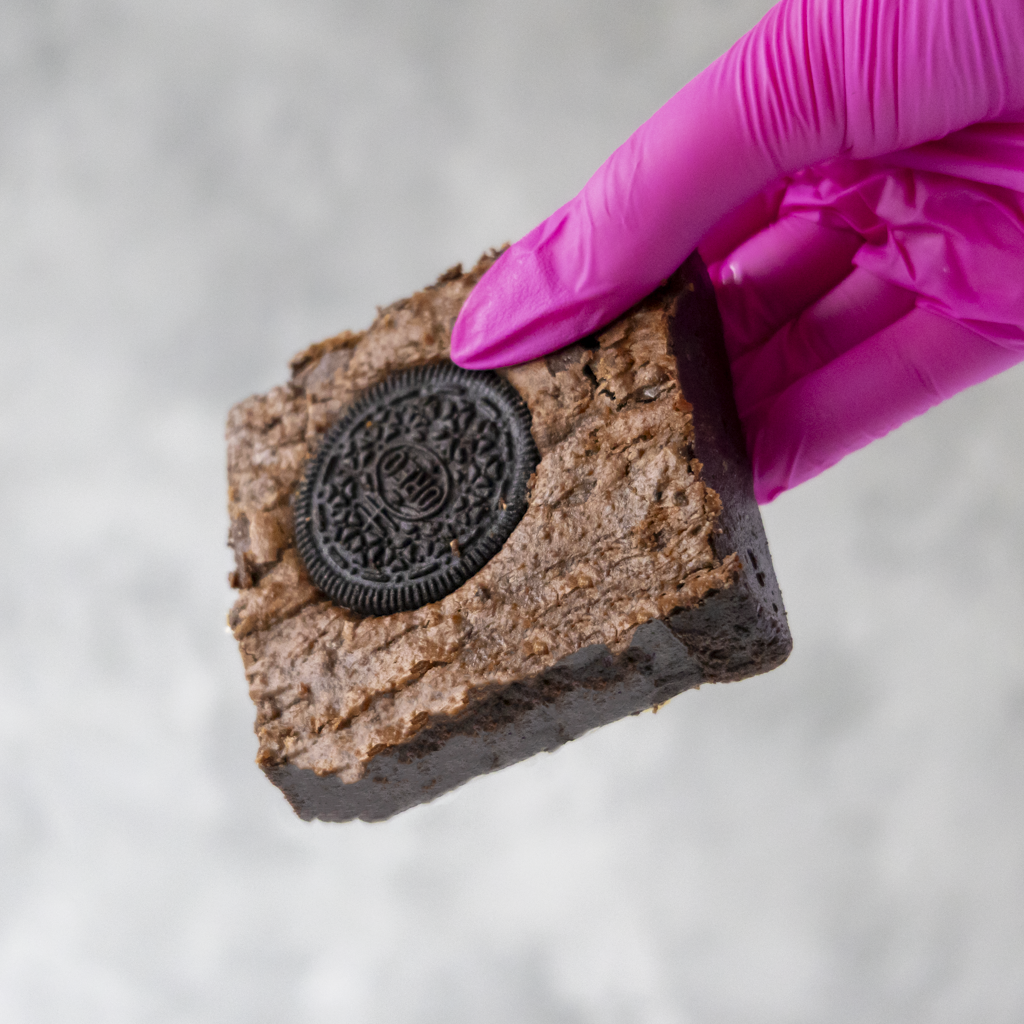 Cookies 'n Cream Brownie Slab by DoughGirl UK with an Oreo cookie in a hand wearing a pink glove on a light gray background