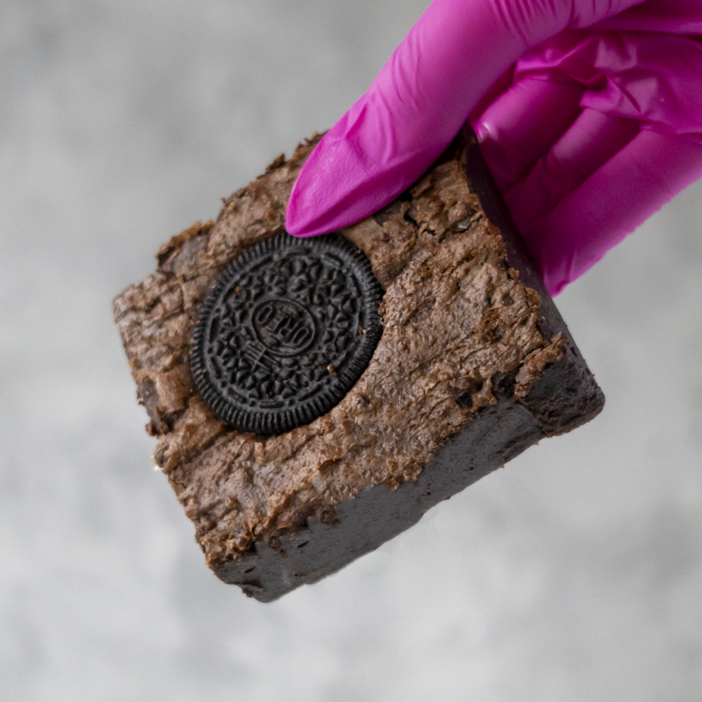 DoughGirl Cookies 'n Cream Brownie Slab with an Oreo cookie in a hand wearing a pink glove against a neutral background