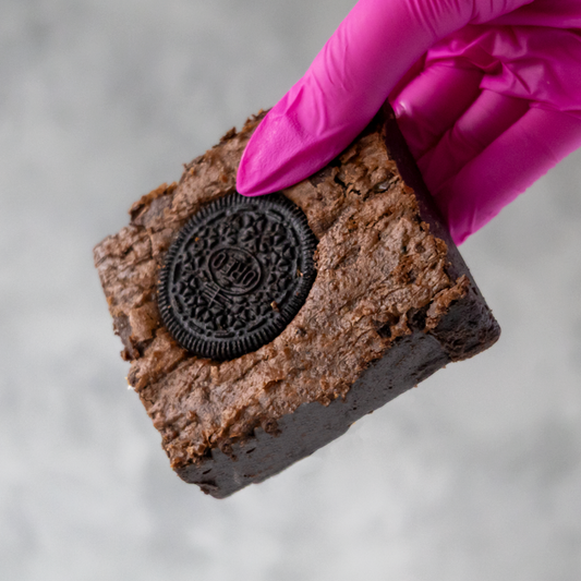 DoughGirl Cookies 'n Cream Brownie Slab with an Oreo cookie in a hand wearing a pink glove against a neutral background