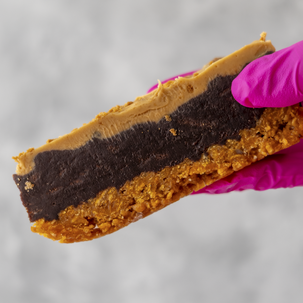 Close-up of a Cornflake Brownie Biscoff Pie Slice with a pink glove holding it, on a gray background