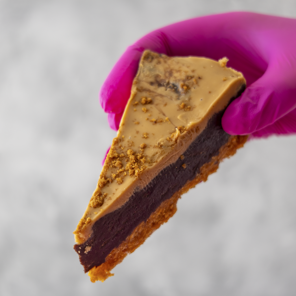 Slice of Cornflake Brownie Biscoff Pie Slice with a chocolate and biscoff spread layer, held by a gloved hand against a neutral background.
