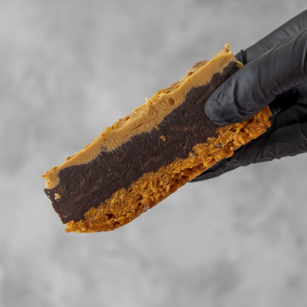 Hand holding a Cornflake Brownie Biscoff Pie Slice with chocolate and biscoff spread, against a gray background.